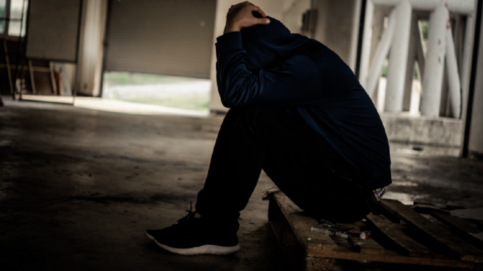 Person sitting on a wooden pallet in a dimly lit room looking down with one hand on their head
